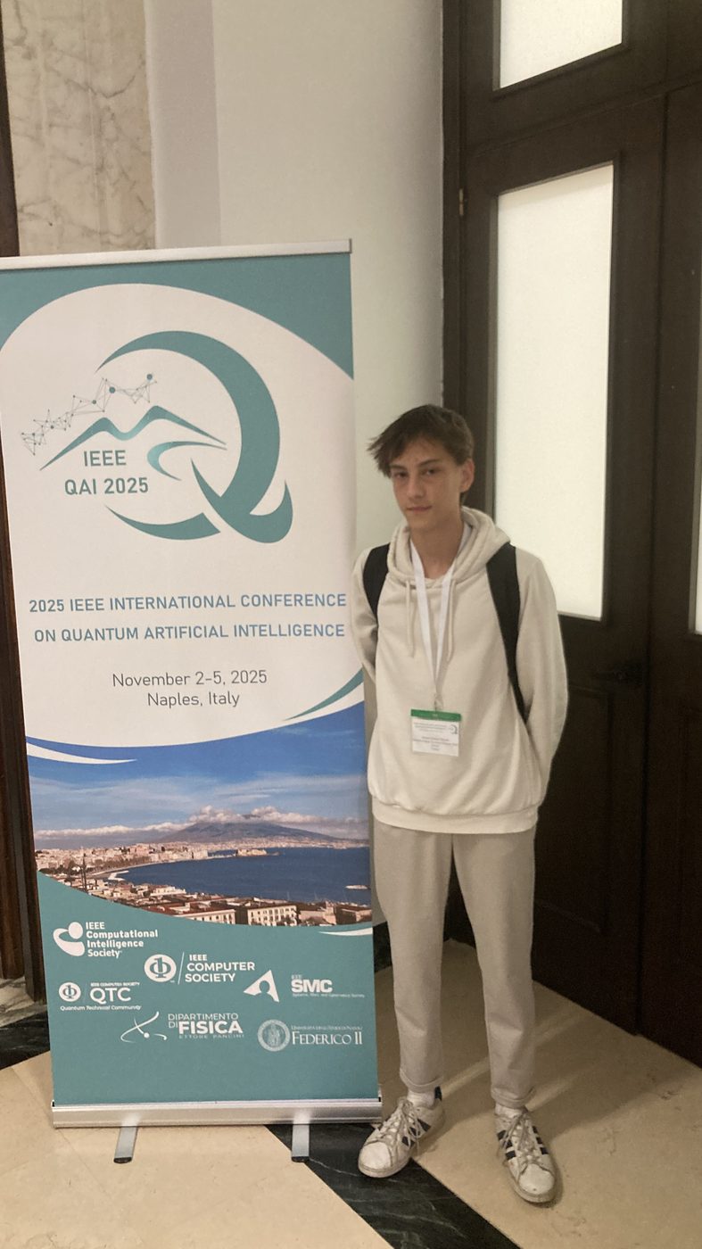 ahmet standing next to the ieee qai 2025 banner in naples